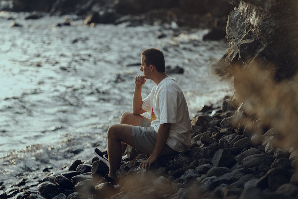 "Llevaba años con ansiedad pensando.." A man sits pensively on a rocky shore by the sea, wearing a white shirt, during the day.