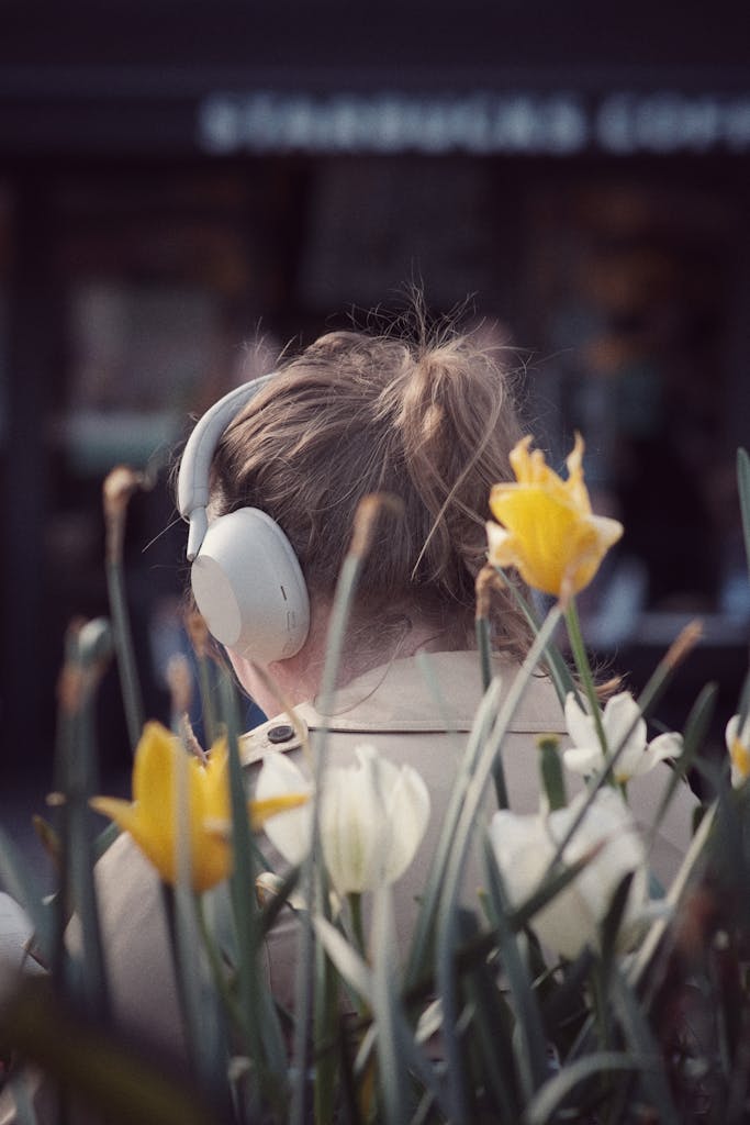 "Sentirme comprendida" A woman enjoys music with headphones amidst spring flowers, city cafe backdrop.