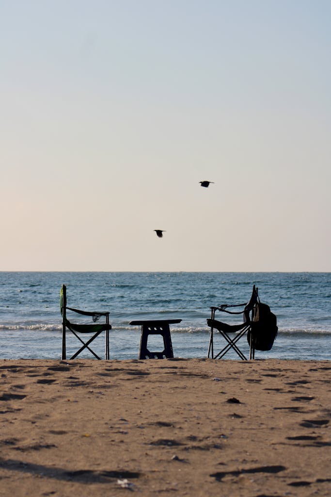 Un lugar en calma Two empty camping chairs and a table on a beach with a serene sea view at sunset.